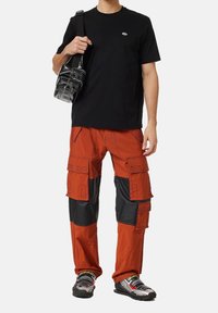 Černé tričko, oranžové a černé cargo kalhoty s náprsními kapsami, vzorované tenisky a malá černá crossbody taška.