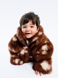 Καφέ ψεύτικη γούνα ζακέτα με λευκά στίγματα, με φερμουάρ και υπερμεγέθη μανίκια, φορεμένη από ένα παιδί σε καθιστή θέση.