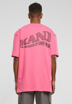 Osoba nosząca jasnoróżowy oversize T-shirt z napisem „KANI THE ORIGINATOR OF STREETWEAR” na plecach, szare spodnie typu cargo.