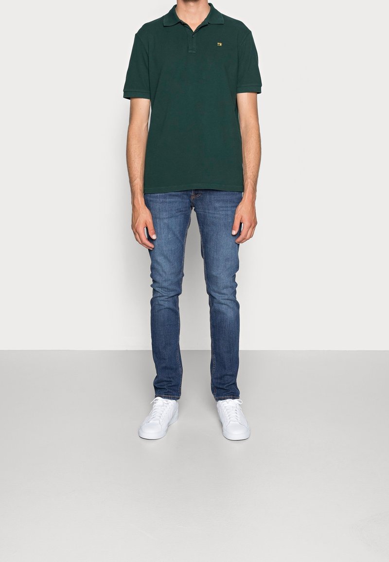 Polo noir avec un col, associé à un jean bleu slim et des baskets blanches. Le tissu semble lisse avec un design classique.