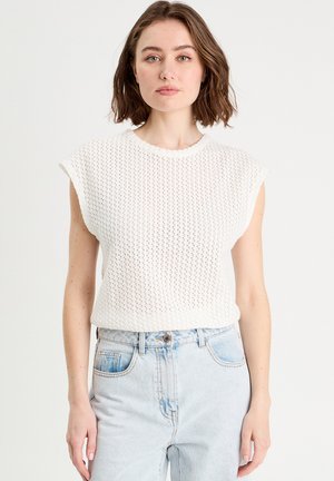 Mujer con cabello corto castaño, vestida con un top sin mangas de punto blanco y jeans de talle alto azul claro, posando frente a un fondo liso.