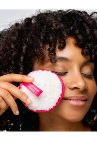 Almohadilla redonda y suave para desmaquillar con una superficie blanca y un borde rosa brillante. La etiqueta dice "REMOVE" en texto blanco. Cabello rizado en el fondo.