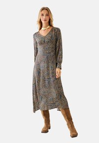 Vestido de manga larga en azul oscuro con patrones de hojas multicolores, con un profundo escote en V y una silueta fluida, combinado con botas marrones hasta la rodilla.