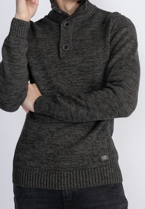 Mand iført mørkegrå strikket sweater med to knapper på høj krave, armene krydset, poserer mod ensfarvet baggrund.