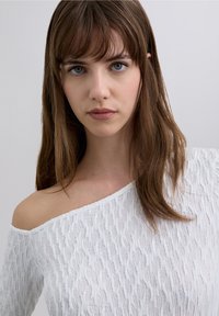 Vit off-shoulder-topp med ett texturerat, vågigt mönster och mjukt tyg. Halsringningen är bred och avslöjar axeln.