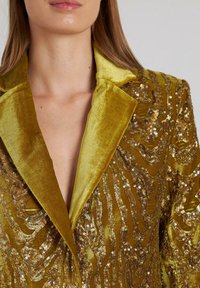 Olijfgroene fluwelen blazer met een gestructureerd oppervlak, versierd met pailletten in golvende patronen. Beschikt over een geknipt kraagje en een diepe V-hals.