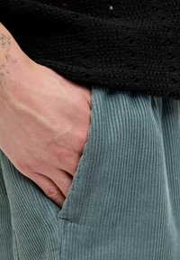 Pantaloni din corduroy de culoare albastru deschis, cu o suprafață texturată, având un buzunar lateral în care se odihnește o mână. Partea de sus este dintr-o țesătură neagră tricotată.
