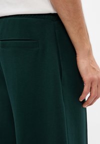 Sötétzöld puha szövetből készült jogger nadrág oldalzsebbel és rugalmas derékkal, sima textúrával és letisztult vonalakkal.