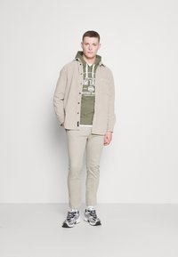 Light beige button-up shirt over olive green hoodie, cream-colored pants, and black and silver sneakers. Simple design, casual fit.