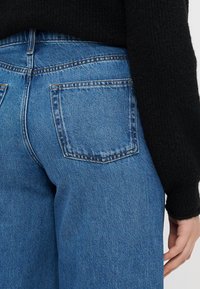 Jeans din denim albastru au un design cu talie înaltă, picioare drepte și două buzunare posteriori. Materialul are un aspect clasic, texturat.