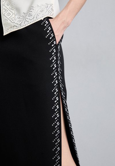 Fustă neagră cu o deschidere laterală, împodobită cu un model geometric de capse argintii pe margine, purtată împreună cu un top alb texturat.
