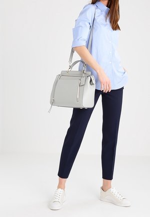 Kobieta w jasnoniebieskiej koszuli, granatowych spodniach, białych trampkach, trzymająca szaro-torebkę z paskiem na ramię na jednolitym tle.