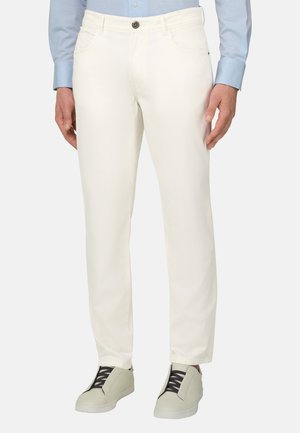 Homme portant un pantalon blanc droit, une chemise bleu clair à manches longues et des baskets blanches avec des lacets noirs, debout devant un fond uni.