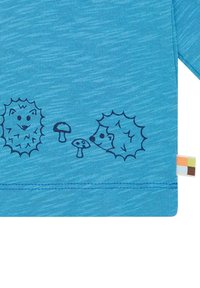 Helleblaue Baumwollbluse mit zwei Igeln und Pilzen im dunkelblauen Druck am unteren Ende. Enthält ein mehrfarbiges Etikett an der Seite.