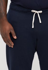 Granatowe dresy z troczkiem w pasie, charakteryzujące się gładką teksturą i elastycznym pasem. Ręka spoczywająca na boku z widocznym odcieniem skóry.