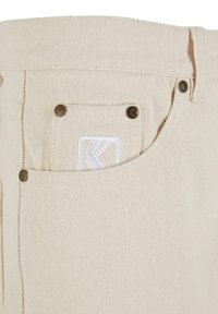 Pantaloni din denim bej cu un buzunar dorsal vizibil, prezentând un logo alb brodat. Nasturii din metal accentuează buzunarul.