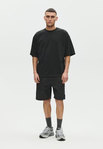 Jeune homme debout, face à l'avant, portant un t-shirt noir ample, un short cargo noir, des chaussettes gris clair montantes et des chaussures de course grises.