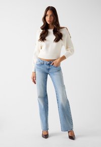 Biały krótki sweter z prążkowanym wzorem, połączony z jasnoniebieskimi jeansami o prostych nogawkach. Modelka ma na sobie brązowe szpilki z zaostrzonym noskiem.