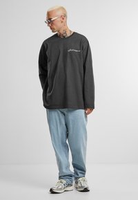 Langærmet grå skjorte med hvid broderet tekst, parret med lyseblå jeans og sølvfarvede sportssko. Modellen står og ser til siden.