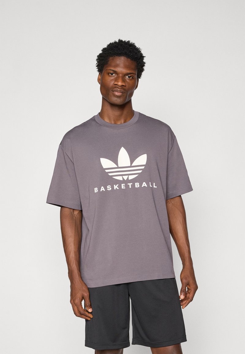 Pilkas medvilnės marškinėliai su dideliu baltu Adidas logotipu, kuriame yra trys lapai ir žodis "KREPŠINIS" žemiau. Trumpomis rankovėmis ir laisvu kirpimu.