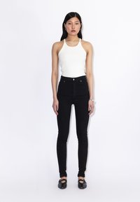 Młoda kobieta z długimi ciemnymi włosami, ubrana w biały top bez rękawów, czarne skinny jeansy z wysokim stanem, czarne płaskie buty i srebrną biżuterię.