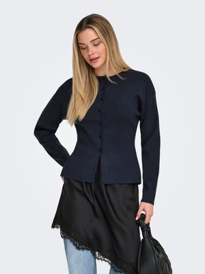 Sieviete ar gariem blondiem matiem, valkājot tumši zilu pogājamu džemperi, melnu mežģīņu apdari apvilktu svārku virs zilām džinsu biksēm, turot melnu rokas somu.