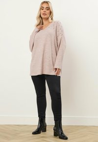 Vaaleanpunainen pitkähihainen neulepaita, jossa on V-pääntie ja koristellut olkapäät, yhdistetty mustiin tiukkoihin housuihin ja kiiltäviin mustiin nilkkasaapikkaisiin.