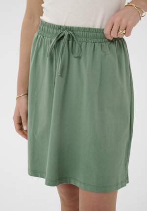 Nő állítja a zsinóros derekú, térdig érő zöld szoknyát, fehér felsőt és arany ékszereket visel a kezén.