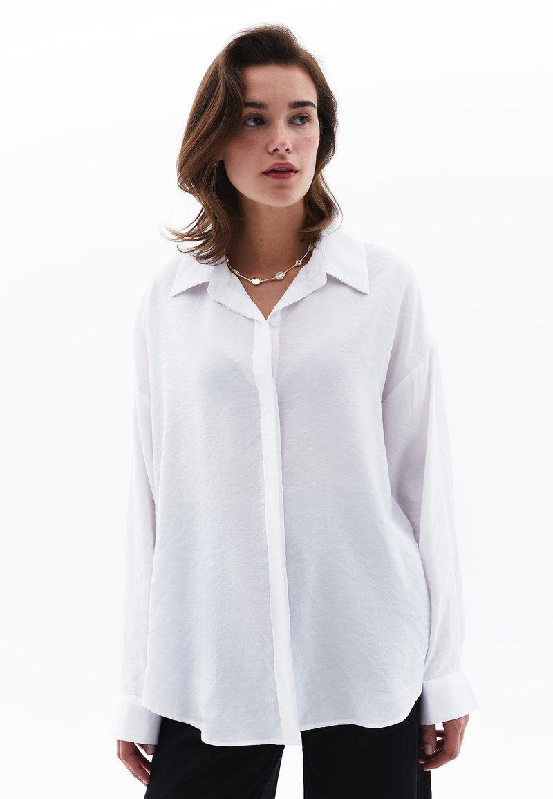 Camisa blanca de manga larga con cuello, con un frente abotonado, corte holgado y tejido texturizado; combinada con pantalones negros.
