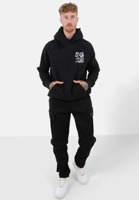 Sudadera negra con estampado gráfico en el lado izquierdo del pecho, combinada con pantalones cargo negros. Zapatillas blancas completan el atuendo.