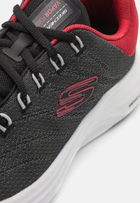 Scarpa sportiva nera con tomaia intrecciata testurizzata, accenti rossi, lacci grigi e suola bianca con un logo prominente sul lato.