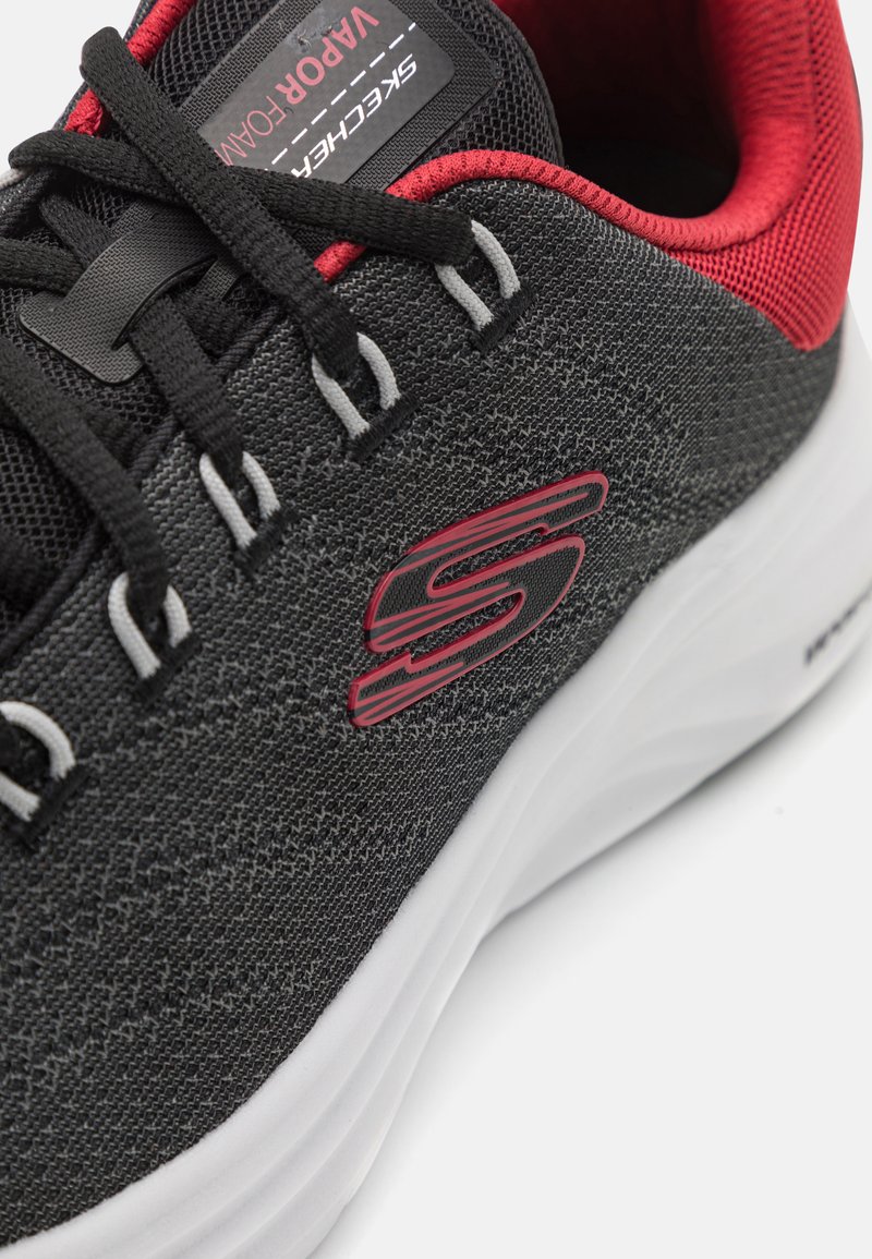 Zapatilla deportiva negra con parte superior de tejido texturizado, acentos rojos, cordones grises y una suela blanca con un logotipo prominente en el lateral.