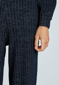 Höger hand vilar vid låret, iförd mörka, strukturerade tygbyxor och matchande långärmad topp mot en ljus bakgrund.