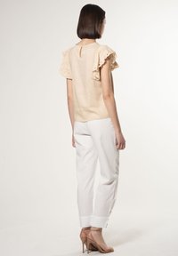 Blusa beige con maniche in pizzo a volant, caratterizzata da un dettaglio a chiave sul retro, abbinata a pantaloni bianchi e sandali con tacco nudo.