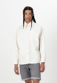 Camicia crema con bottoni e tasca sul lato sinistro del petto, maniche lunghe arrotolate ai polsi, abbinata a pantaloni corti in denim grigi. Texture del tessuto liscia.