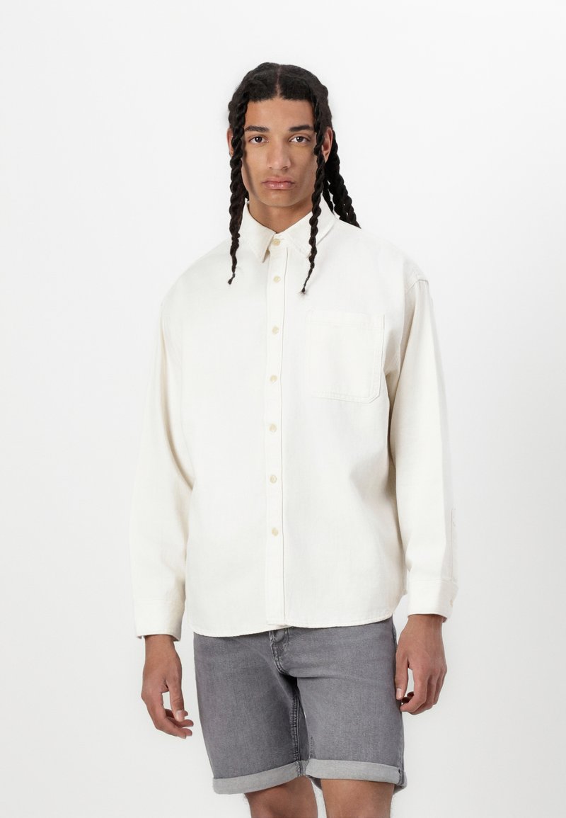Camicia crema con bottoni e tasca sul lato sinistro del petto, maniche lunghe arrotolate ai polsi, abbinata a pantaloni corti in denim grigi. Texture del tessuto liscia.