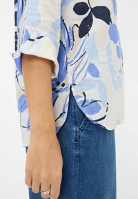 Floral print blouse made of lightweight fabric in various shades of blue and white, featuring rolled sleeves and side slits for breathability.