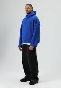Blauwe oversized hoodie met capuchon, gemaakt van zachte stof, gecombineerd met zwarte wijde broekspijpen en stevige zwarte schoenen met gekleurde veters.