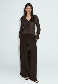 Blouse brune transparente avec un col volé et des manches longues, associée à un pantalon ample marron. Le tissu semble léger et texturé.