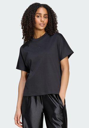 Kvinde med krøllet hår iført en sort Adidas t-shirt og skinnende sorte Adidas bukser, stående med den ene hånd i lommen.