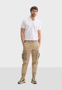 Bílá polo košile se zelenými detaily, béžové cargo kalhoty s bočními kapsami a vícebarevné sportovní boty. Neformální střih a uvolněný styl.