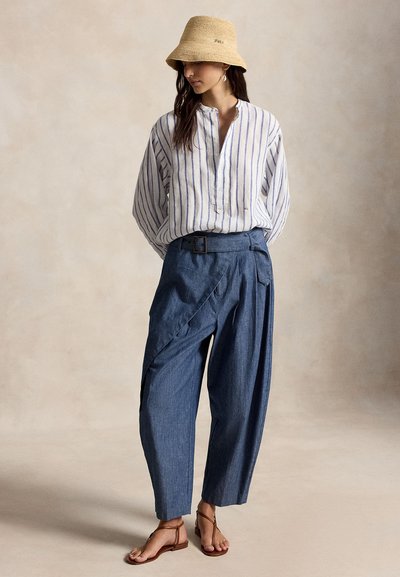 Kvinne iført beige vevd bøttehatt, hvit og blåstripet løs skjorte, blå vidbukser med høy midje og belte, og brune sandaler, stående.
