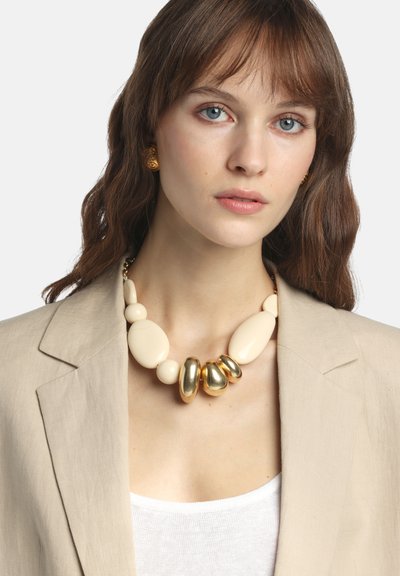 Vastag nyaklánc krémszínű és arany tónusú gyöngyökkel, sima textúrával, arany láncon. Bézs blézer fölött viselve.