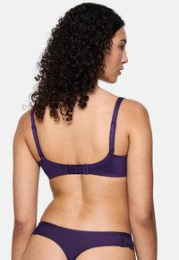 Mulher com cabelo encaracolado a fazer modelo de um soutien roxo e lingerie combinando com detalhes em renda, vista de costas contra um fundo simples.
