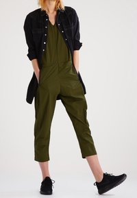 Mono verde con escote en V, dos bolsillos frontales y patas recortadas, sobrecamisado con una camisa de mezclilla negra. Combinado con zapatillas negras.