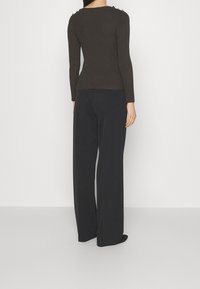 Mujer vestida con una blusa ajustada de manga larga marrón oscuro y pantalones amplios negros de pierna ancha, de pie contra un fondo blanco liso.