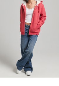 Person wearing a red zip-up hoodie, white lace-trimmed tank top, loose blue jeans, and white sneakers standing with legs crossed.
