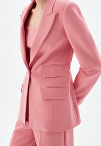 Kobieta w dopasowanej różowej marynarce z klapami i pasujących spodniach, stojąca na tle jednolitego białego tła.