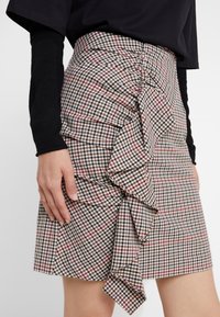 Jupe à carreaux en marron, noir et rouge avec des volants. Fabriquée en tissu au toucher doux. Taille ajustée, longueur genou.