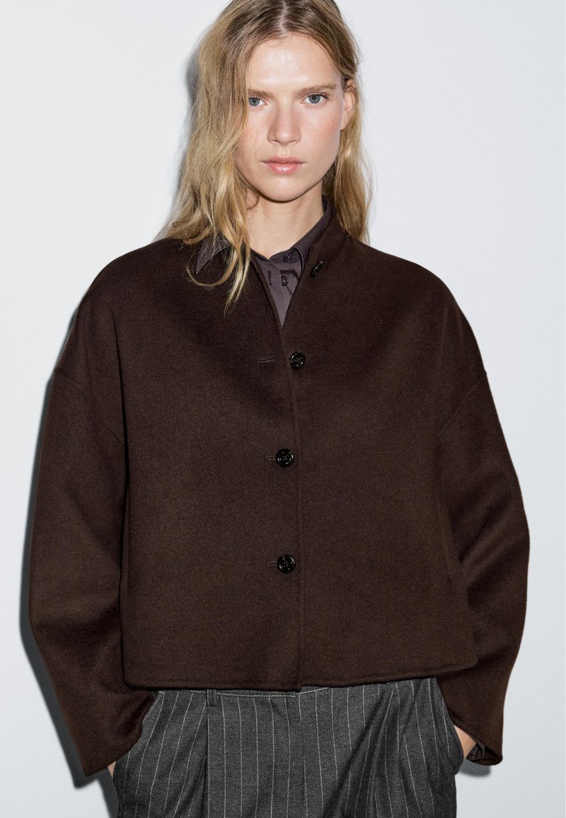 Brown wool cropped jacket with a round neckline, front button closure, and relaxed fit, paired with a dark button-up shirt and pinstriped trousers.